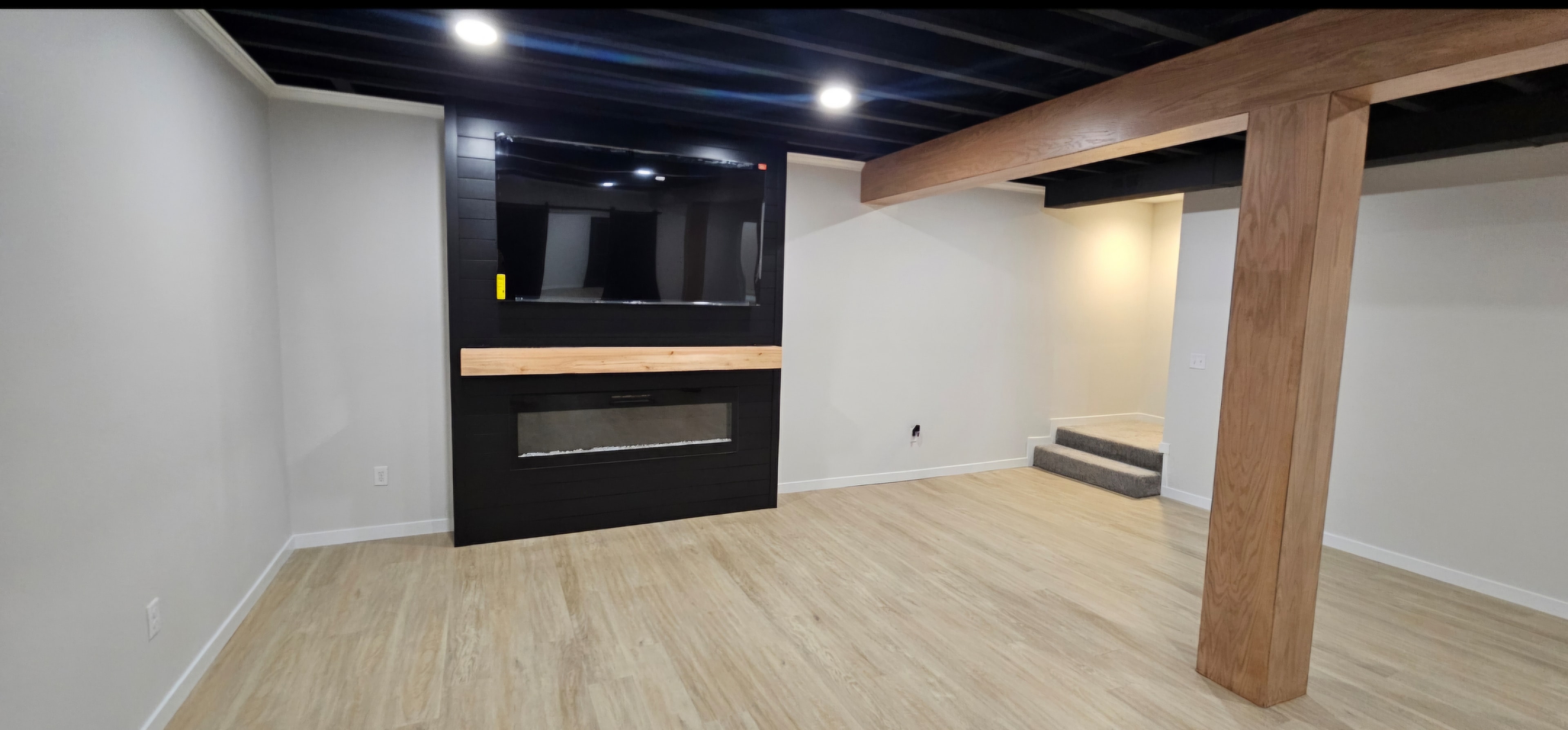 Finished basement living area with black fireplace feature wall, wood mantel and wrapped structural beam.